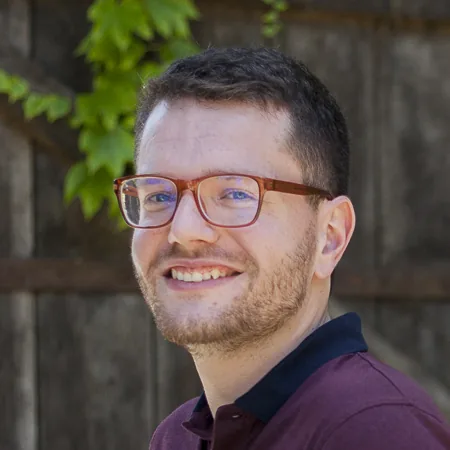 Photographie de Thomas Ghys, souriant, lunettes à monture bordeaux et polo prune, devant une palissade en bois couverte de lierre.